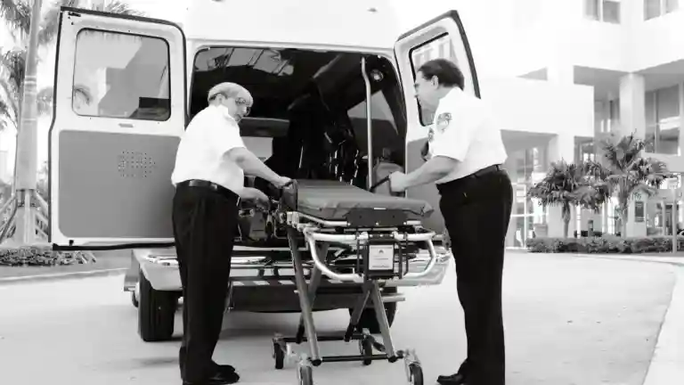 Deux secouristes chargent un brancard à l’arrière d’une ambulance en noir et blanc.