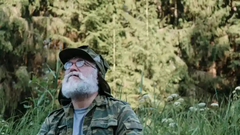 Un homme barbu vêtu d’une veste de camouflage se tient dans un paysage boisé.