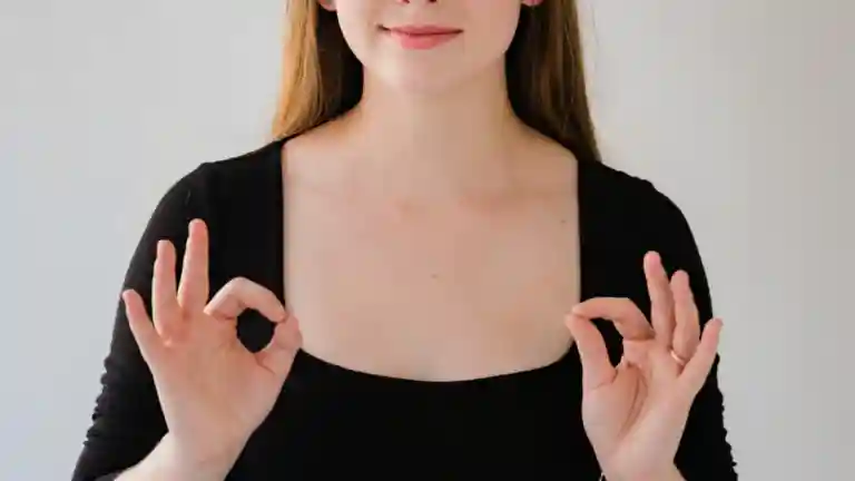 Une femme en tenue noire effectue des gestes de langue des signes avec les deux mains, devant un fond clair.