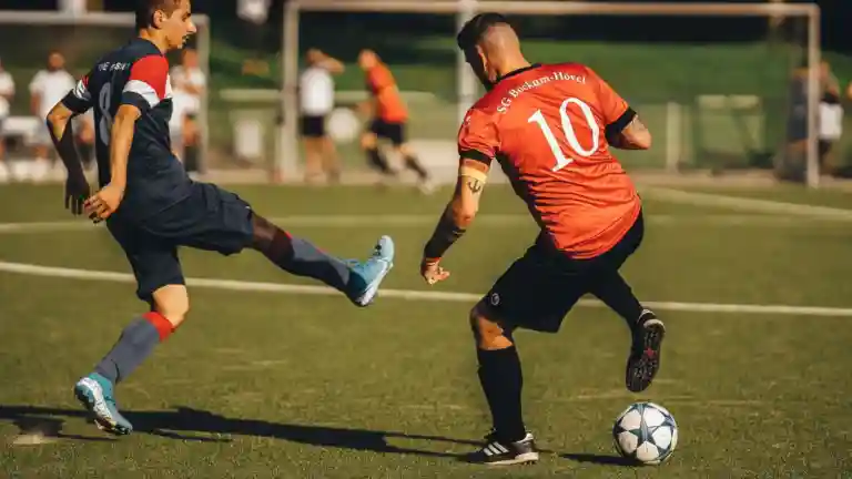 Deux footballeurs disputent un ballon sur un terrain en plein air, l'un portant un maillot bleu et l'autre orange
