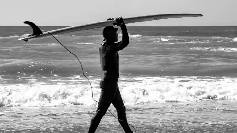 Surfeur portant un longboard au bord de l’océan, en noir et blanc