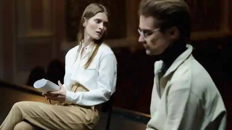 Une comédienne vêtue d'une chemise claire échange avec un acteur en tenant un texte sur papier